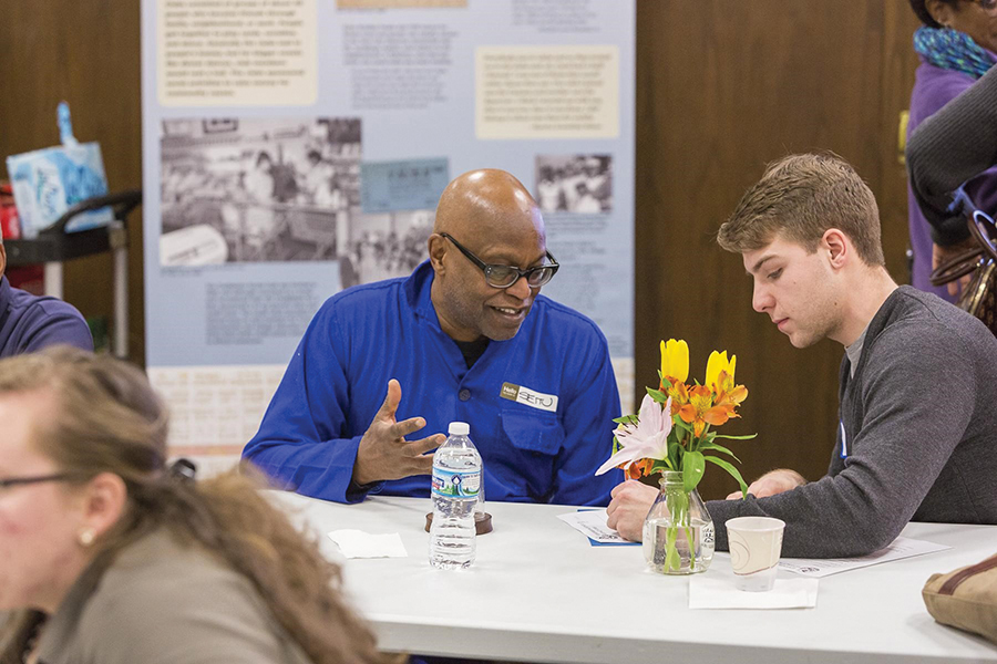 In a crowded room, the camera zooms in on a History Harvest student who is going over paperwork with local artist, Seitu Jones. There are more placards of Rondo’s history along the walls in the background.