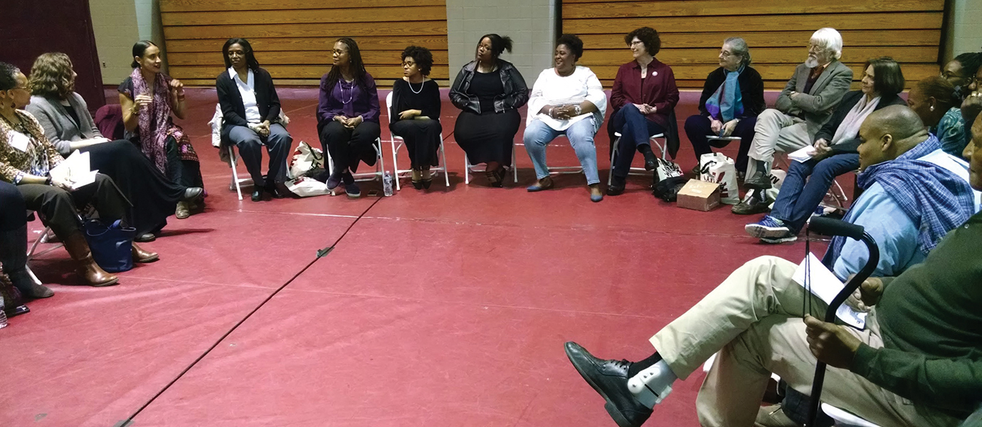 Activists of all ages sit in a circle of chairs listening to a woman speaking.