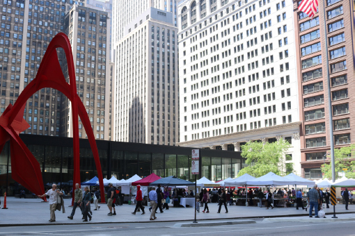 A large sculpture appears to the left of the market stalls. Large office buildings are in the background.