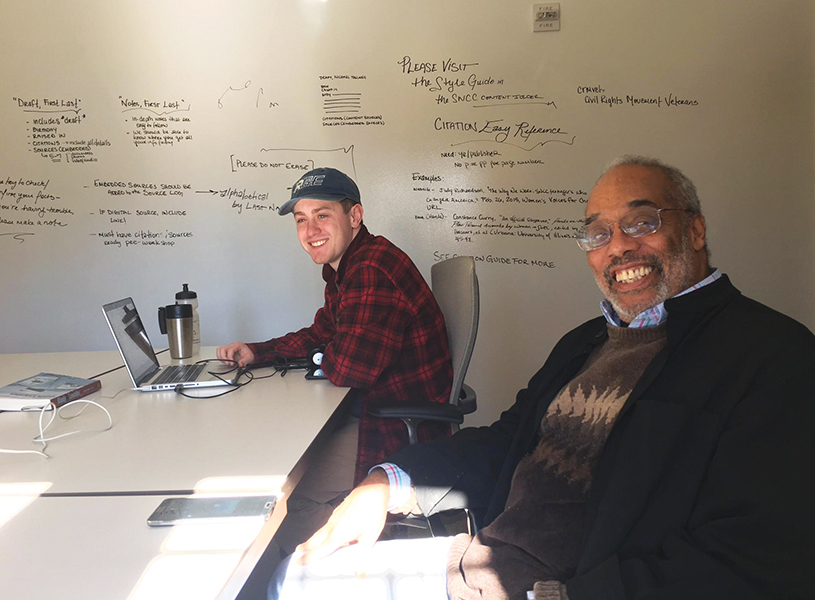 Charlie and Todd sit on the same side of the table, smiling, in the SNCC Digital Gateway project room. Todd has his computer in front of him, and Charlie is leaning back in his chair.