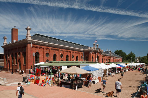 In front of a large brick building, stalls are lined up. Several people are milling around.