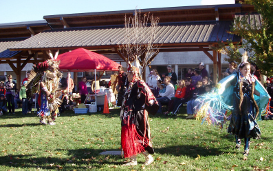 Native Americans dressed in traditional attire performing dances on a green space at the market. A pavilion and a small pop-up tent are behind them.