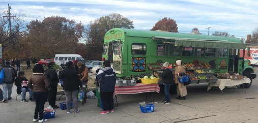 People crowd behind a green school bus with produce stands and stalls lining the outside of the bus. This bus was converted into a mobile market.